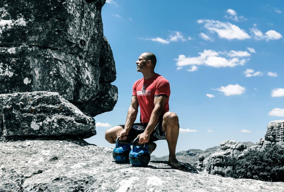 A man exercising with kettlebells on rocky ground under a blue sky