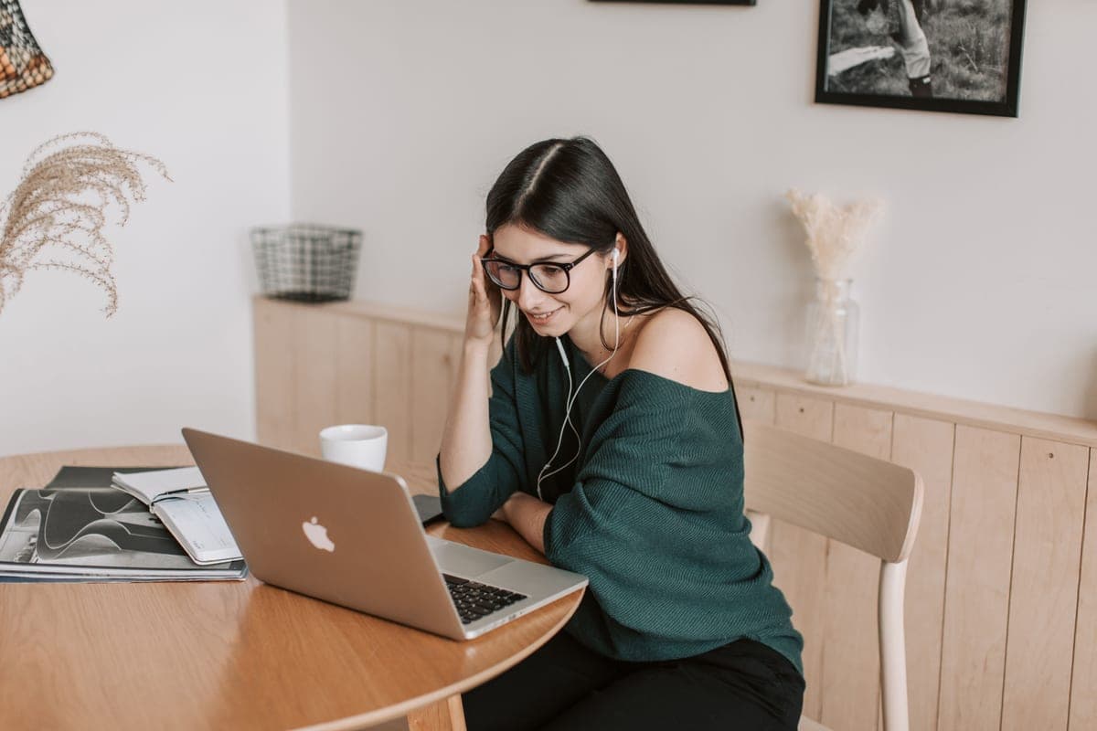 A young woman in eyeglasses listening to music on earbuds at a laptop
