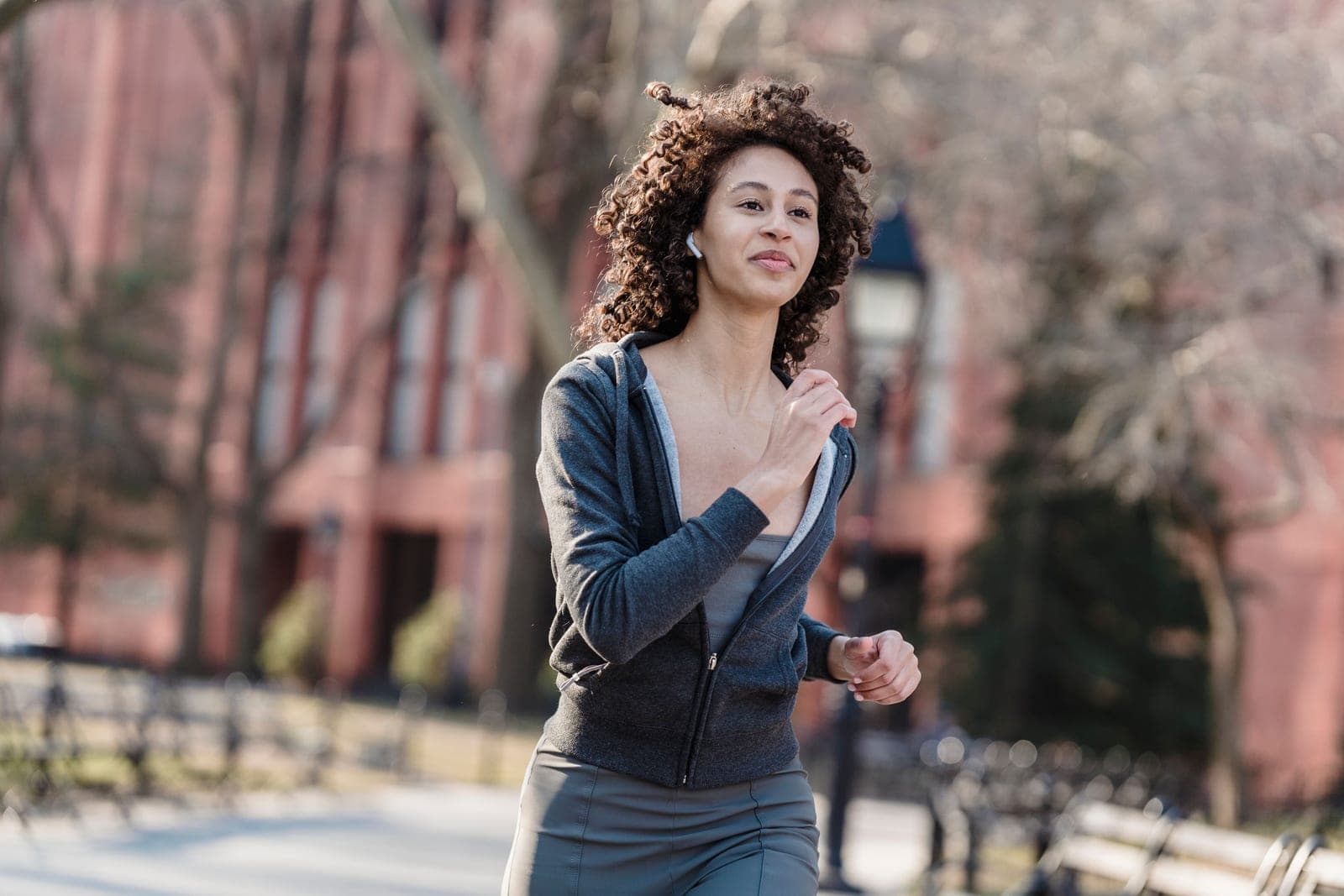 A curly-haired woman in activewear jogging through a park with wireless earbuds in